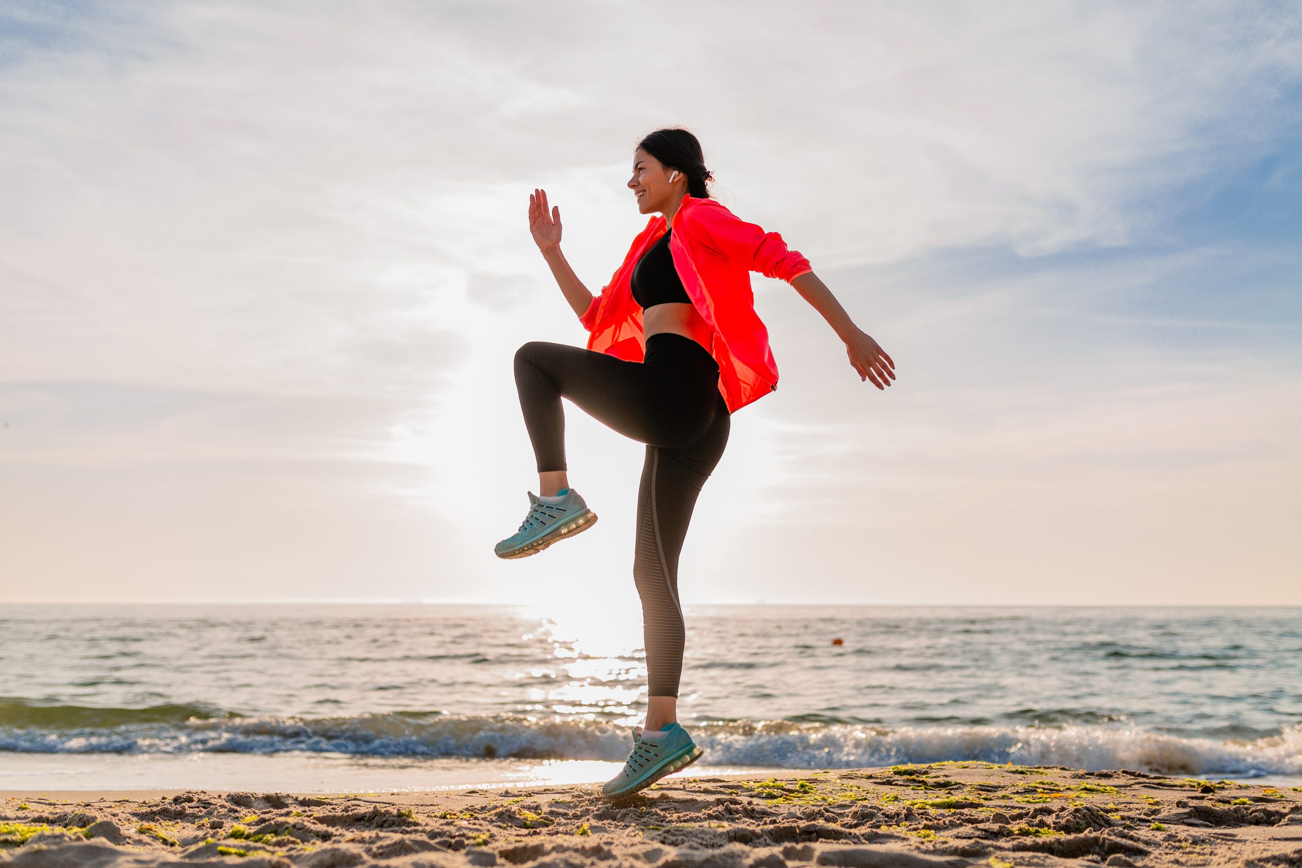 young smiling attractive slim woman doing sports in morning sunrise jumping on sea beach in sports wear, healthy lifestyle, listening to music on earphones, wearing pink windbreaker jacket, having fun